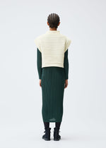 A person with braided hair faces away, wearing the CREPE KNIT by PLEATS PLEASE ISSEY MIYAKE over a dark green pleated dress and black ankle boots, posed against a plain light background.