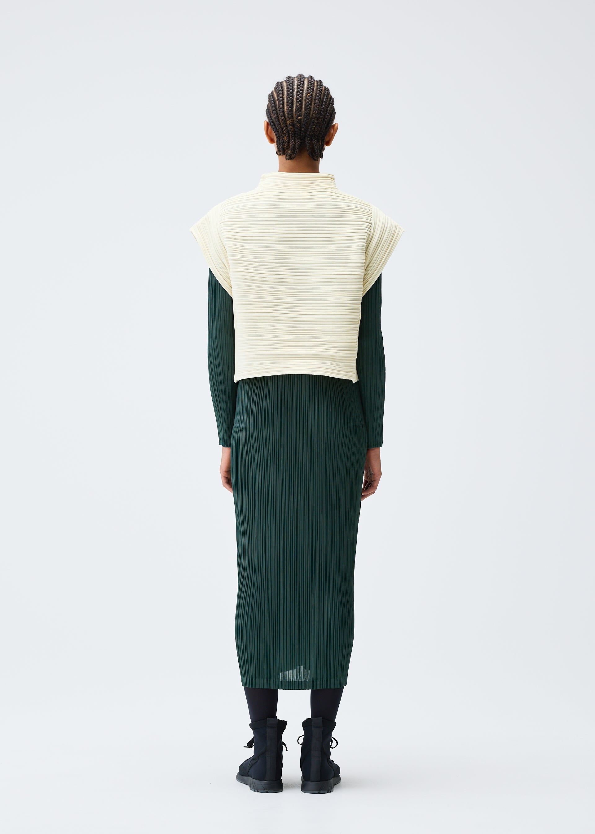 Zoomed view A person with braided hair faces away, wearing the CREPE KNIT by PLEATS PLEASE ISSEY MIYAKE over a dark green pleated dress and black ankle boots, posed against a plain light background.