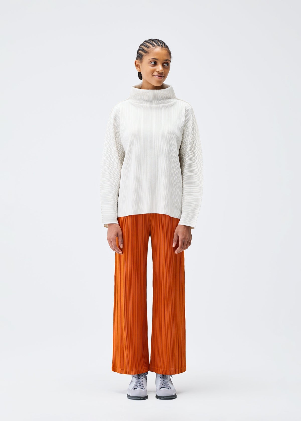A person stands facing forward against a white background, wearing the PLEATS PLEASE ISSEY MIYAKE A-POC SHELL white high-neck long-sleeve top, orange wide-leg pleated pants, and gray sneakers. view 5 of 8