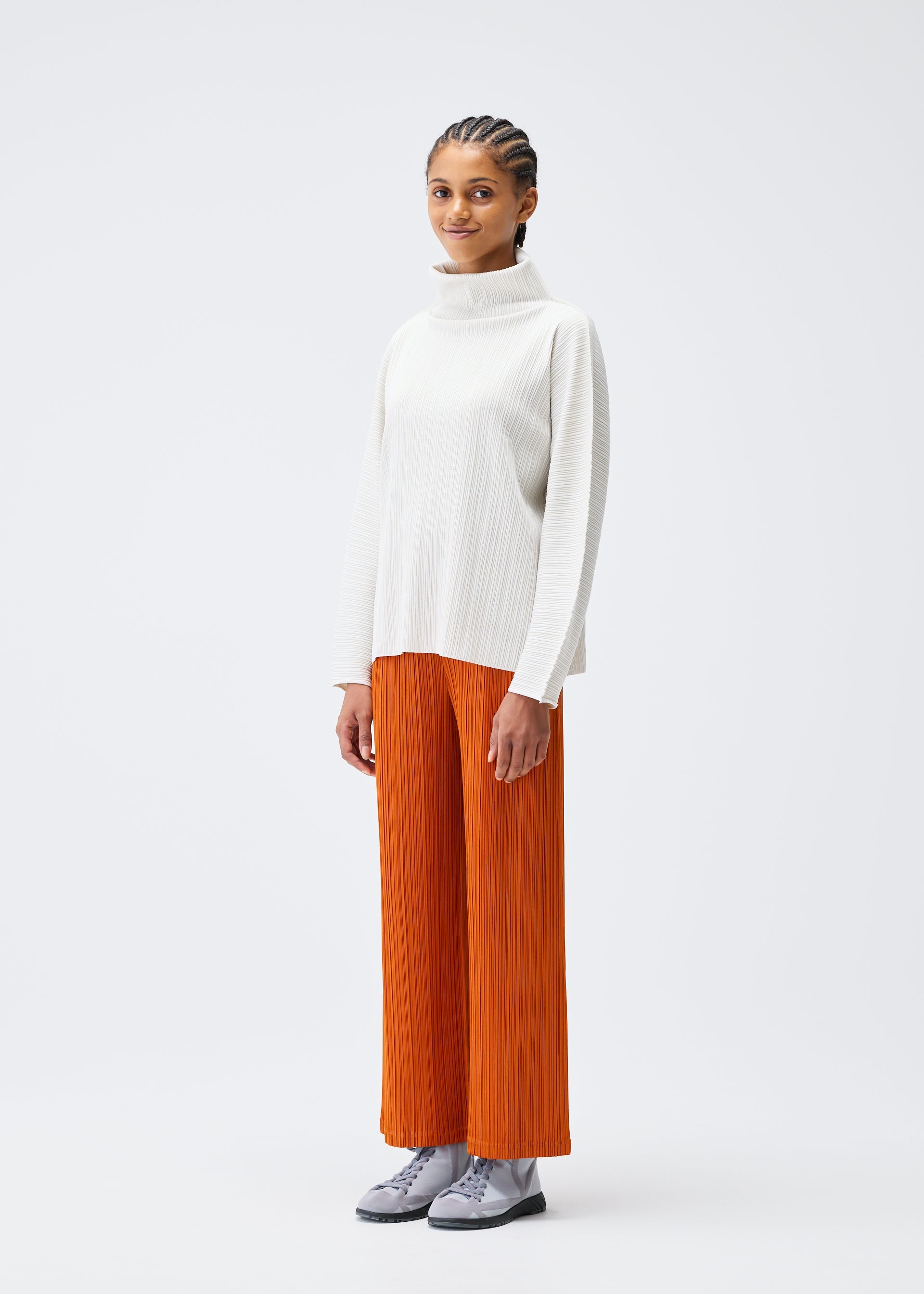 Zoomed view A person with braided hair and a calm expression stands against a white background, wearing PLEATS PLEASE ISSEY MIYAKE’s A-POC SHELL high-neck top and orange pleated wide-leg pants with gray shoes.