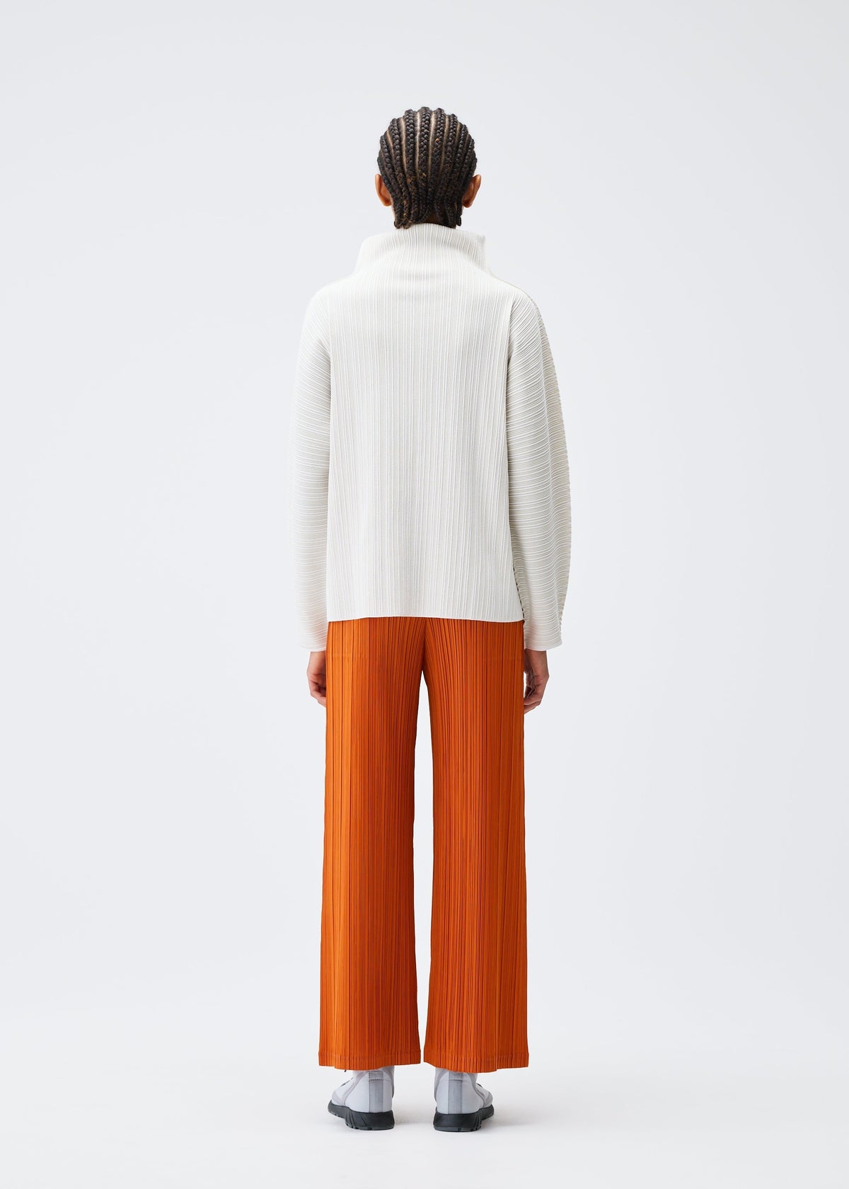 A person with braided hair faces away, wearing the PLEATS PLEASE ISSEY MIYAKE A-POC SHELL white high-neck top with stretchy long sleeves and loose orange pleated pants, paired with light gray socks and dark sandals against a plain white background. view 7 of 8