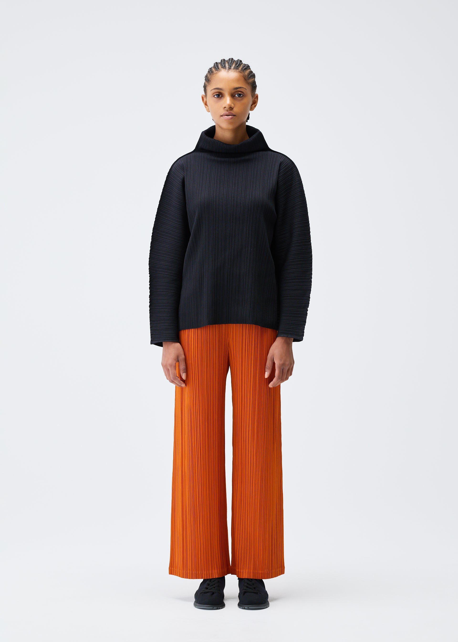 Zoomed view A person stands against a plain white background wearing the A-POC SHELL turtleneck sweater and bright orange wide-leg pleated pants by PLEATS PLEASE ISSEY MIYAKE, paired with black shoes, looking straight ahead with a neutral expression.