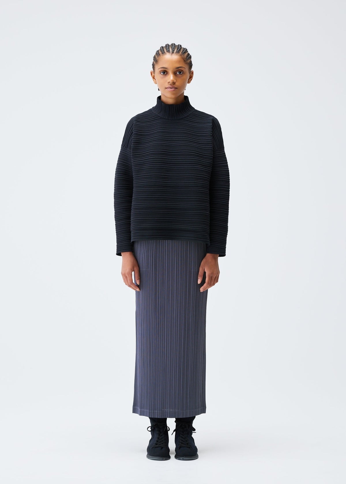A person stands against a plain white background, wearing the PLEATS PLEASE ISSEY MIYAKE CREPE KNIT high-neck turtleneck sweater and a long pleated gray skirt, with black shoes, looking at the camera with arms at their sides. view 4 of 6