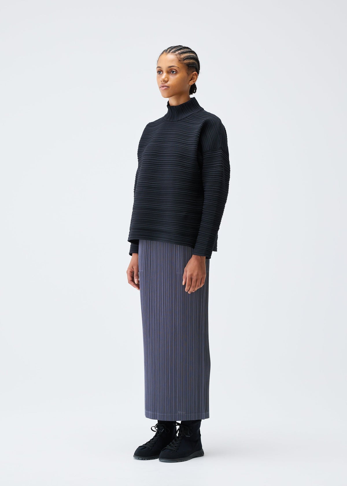 A woman with braided hair stands against a white background, wearing the CREPE KNIT top by PLEATS PLEASE ISSEY MIYAKE, a long gray pleated skirt in recycled polyester, and black boots, facing slightly to the side with a neutral expression. view 5 of 6
