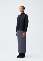 A woman with braided hair stands against a white background, wearing the CREPE KNIT top by PLEATS PLEASE ISSEY MIYAKE, a long gray pleated skirt in recycled polyester, and black boots, facing slightly to the side with a neutral expression.