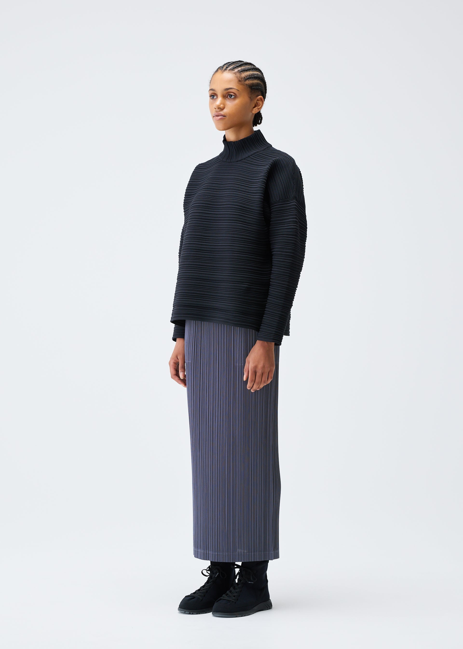 Zoomed view A woman with braided hair stands against a white background, wearing the CREPE KNIT top by PLEATS PLEASE ISSEY MIYAKE, a long gray pleated skirt in recycled polyester, and black boots, facing slightly to the side with a neutral expression.