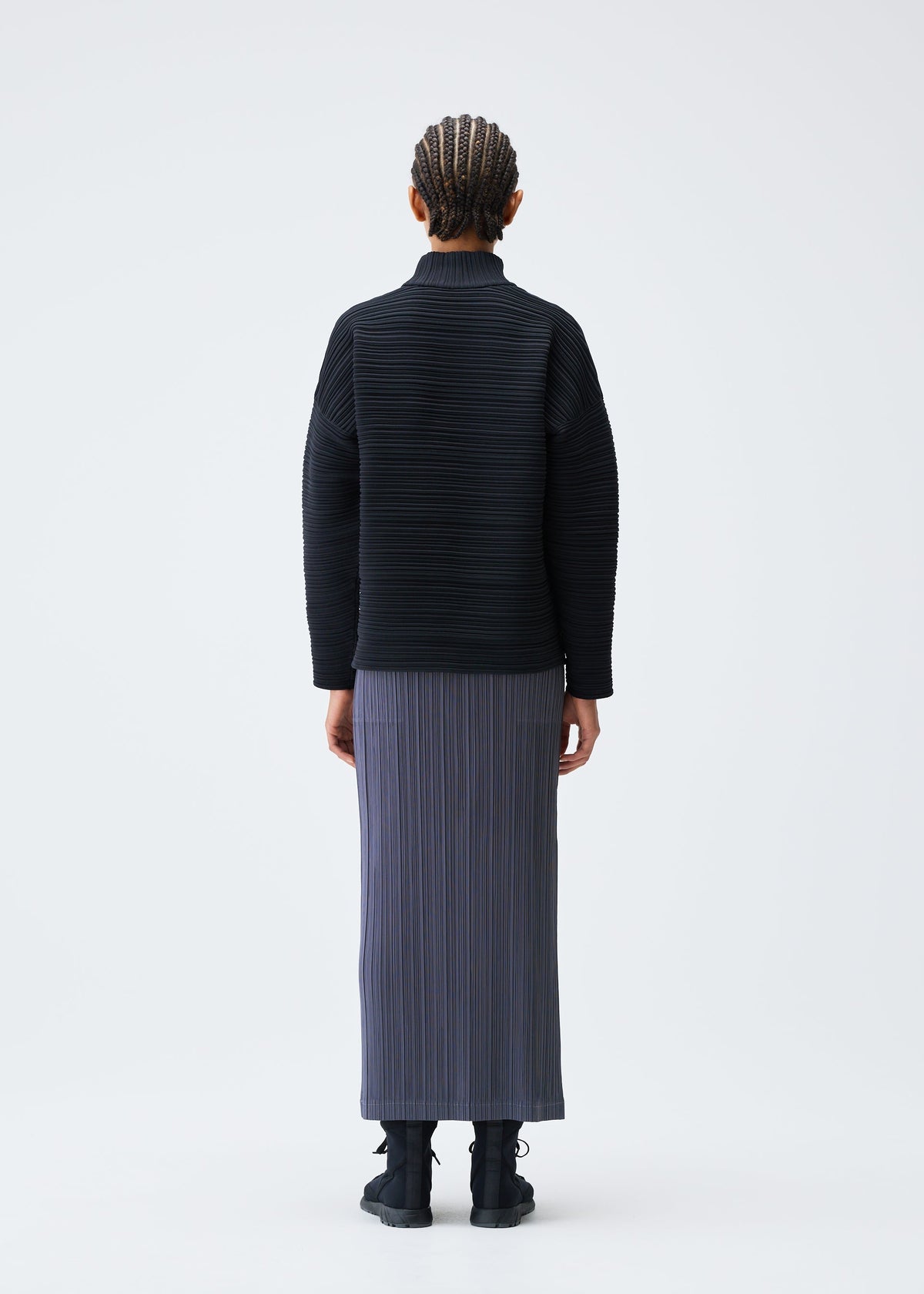 A person with cornrowed hair stands facing away in a PLEATS PLEASE ISSEY MIYAKE CREPE KNIT black high-neck ribbed long-sleeve top, gray pleated skirt, and black shoes against a plain white background. view 6 of 6