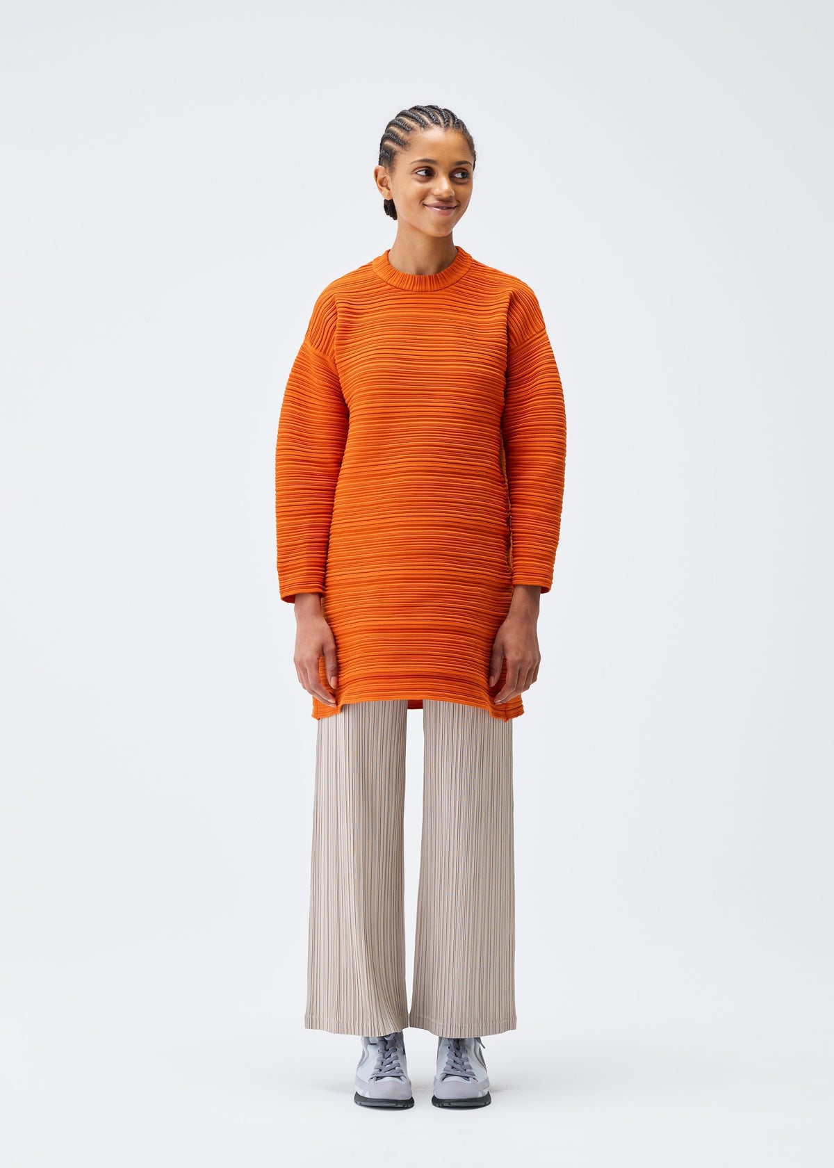 A person stands against a plain white background, smiling gently with hands by their sides. They wear the CREPE KNIT orange tunic and loose beige pleated pants from PLEATS PLEASE ISSEY MIYAKE, paired with gray sneakers. view 4 of 6