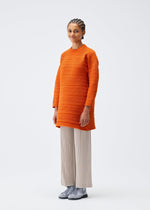 A woman with braided hair smiles, wearing a PLEATS PLEASE ISSEY MIYAKE CREPE KNIT oversized orange sweater, light beige pleated wide-leg pants, and light gray lace-up shoes, standing against a plain white background.