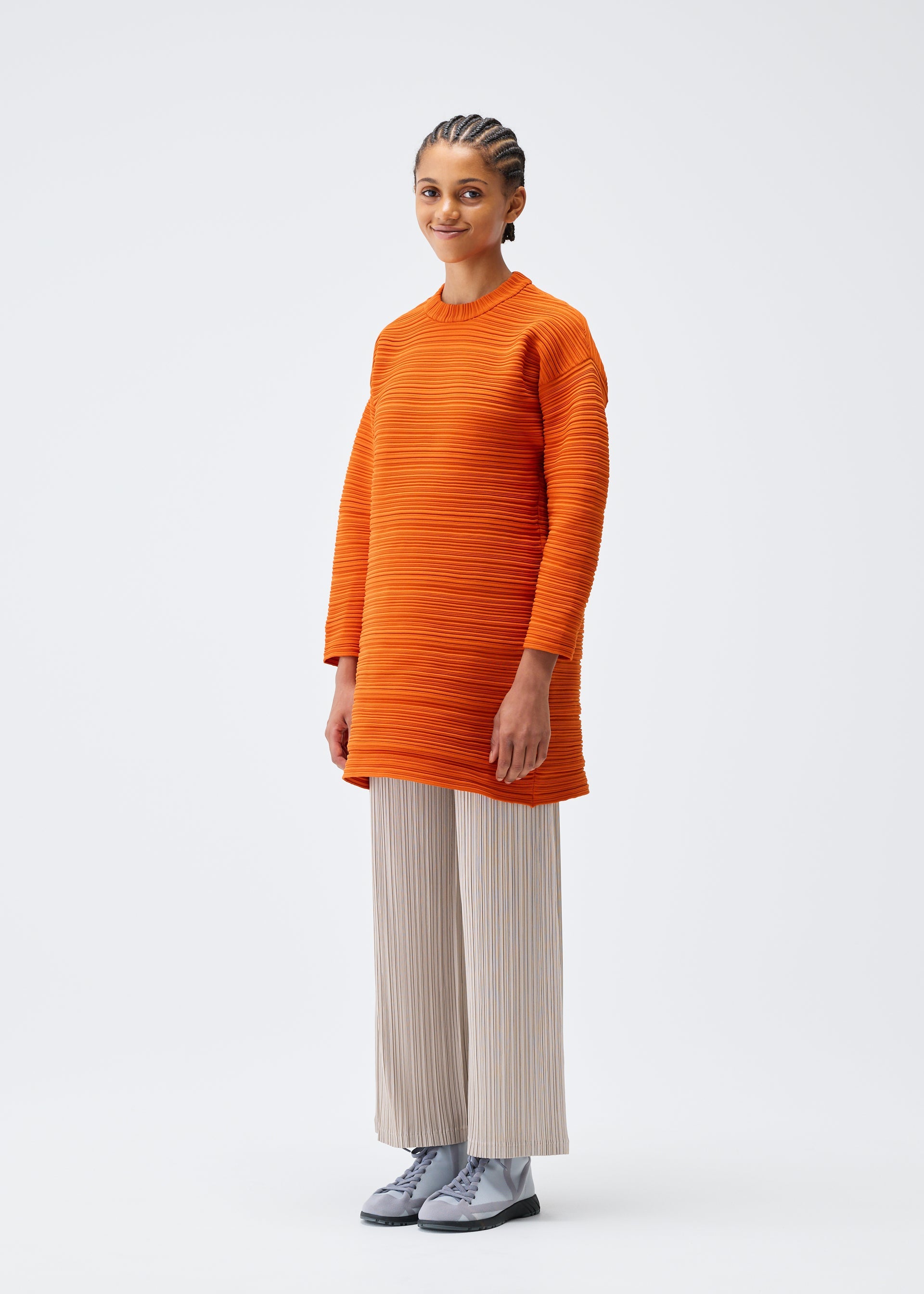 Zoomed view A woman with braided hair smiles, wearing a PLEATS PLEASE ISSEY MIYAKE CREPE KNIT oversized orange sweater, light beige pleated wide-leg pants, and light gray lace-up shoes, standing against a plain white background.