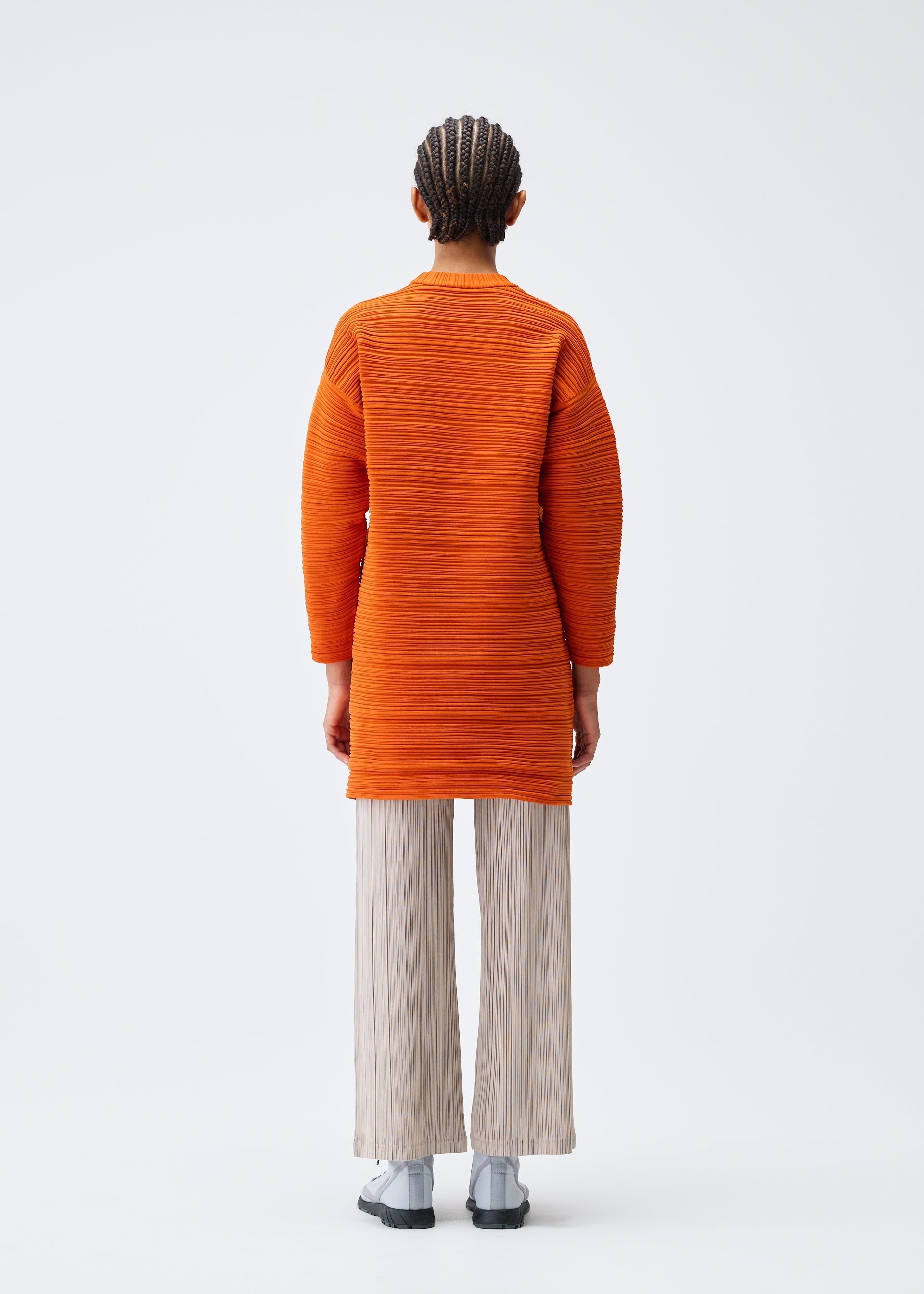Zoomed view A person stands facing away, wearing the PLEATS PLEASE ISSEY MIYAKE CREPE KNIT orange, machine-washable long-sleeved tunic in recycled polyester and light pleated pants with gray shoes against a plain white background.