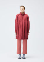 A person faces forward against a plain white background, wearing the PLEATS PLEASE ISSEY MIYAKE PADDED PLEATS COAT in red over pleated light pink pants and gray sneakers.