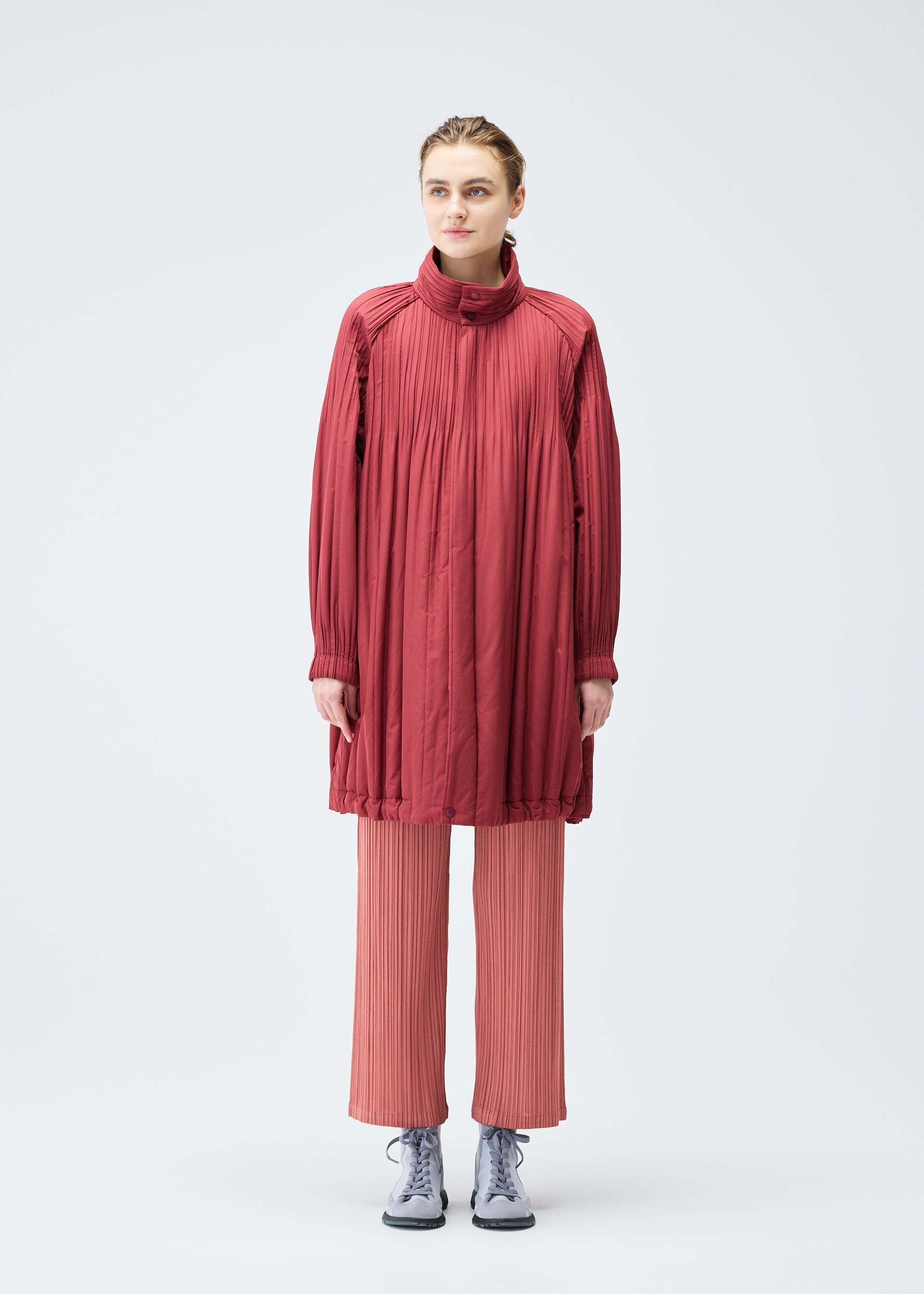Zoomed view A person faces forward against a plain white background, wearing the PLEATS PLEASE ISSEY MIYAKE PADDED PLEATS COAT in red over pleated light pink pants and gray sneakers.