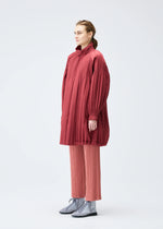 A person stands against a plain background wearing the PLEATS PLEASE ISSEY MIYAKE PADDED PLEATS COAT over wide-legged, pleated pink pants and silver lace-up shoes.
