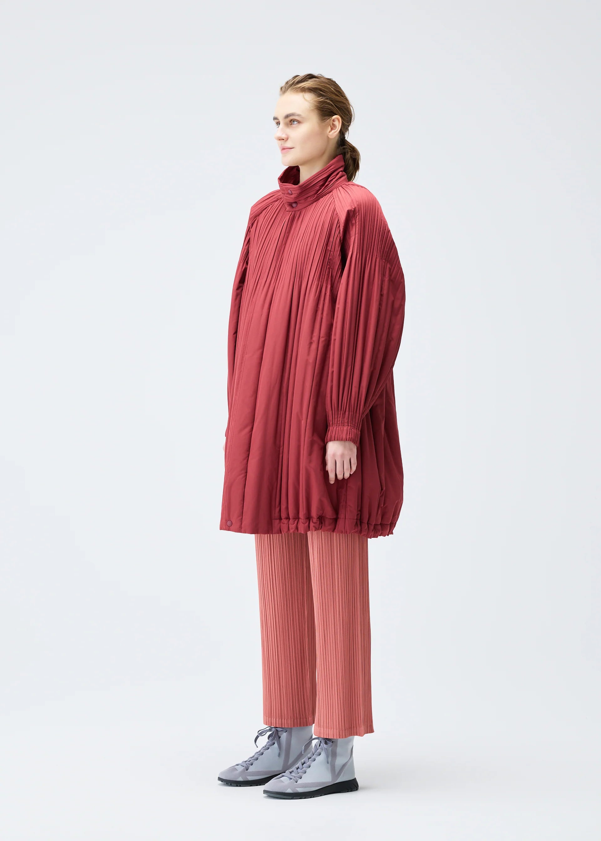Zoomed view A person stands against a plain background wearing the PLEATS PLEASE ISSEY MIYAKE PADDED PLEATS COAT over wide-legged, pleated pink pants and silver lace-up shoes.