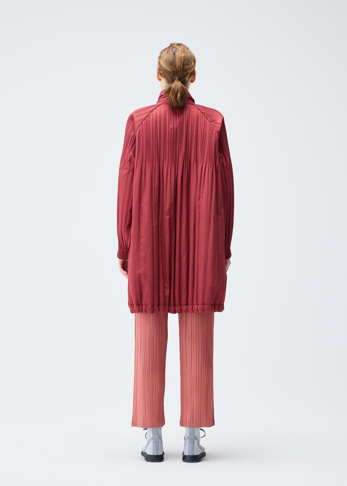A person with light brown hair in a low ponytail faces away, wearing the PADDED PLEATS COAT by PLEATS PLEASE ISSEY MIYAKE over pleated pink pants and black shoes, set against a plain white background. view 5 of 7