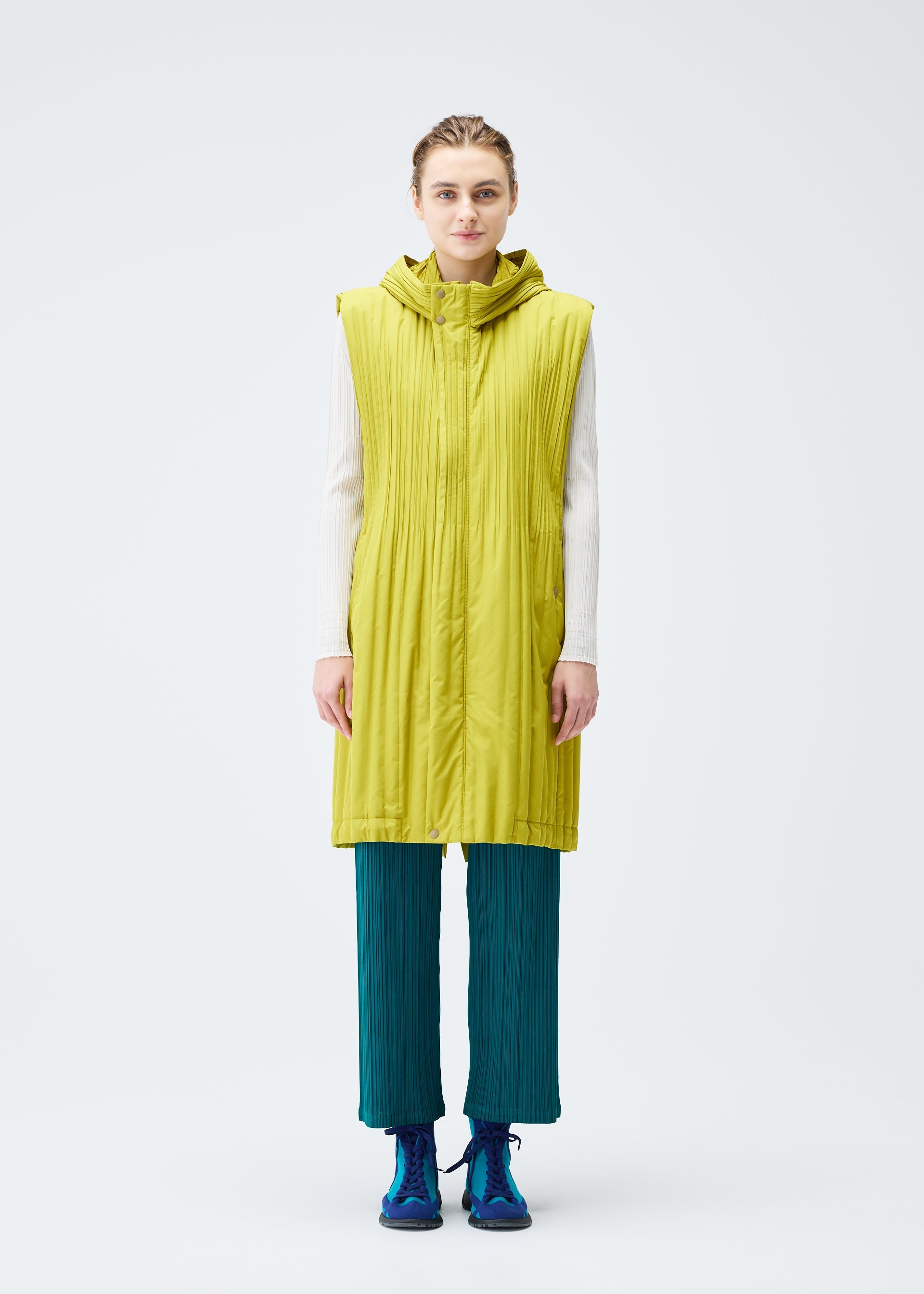 Zoomed view A person faces forward in a PLEATS PLEASE ISSEY MIYAKE PADDED PLEATS COAT—long, sleeveless, lime green, quilted and hooded—layered over a white top, teal Keshi-pleated pants, and blue sneakers against a plain light background.