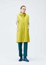 A person stands facing forward in a bright yellow-green PADDED PLEATS COAT by PLEATS PLEASE ISSEY MIYAKE, layered over a white long-sleeve top and teal pleated pants, with blue shoes, set against a plain light backdrop.