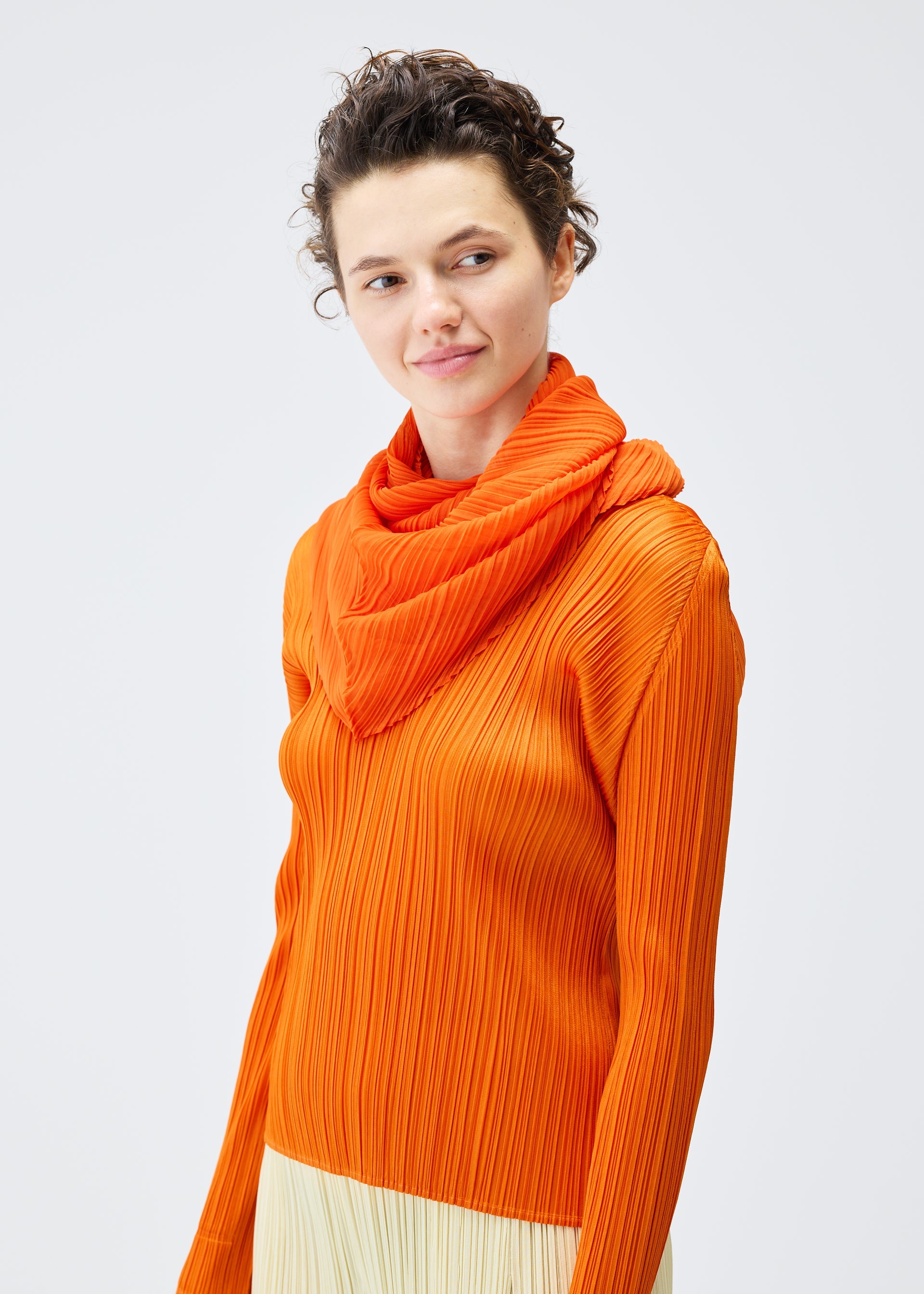 Zoomed view A person with short curly hair wears the bright orange O SCARF DECEMBER by PLEATS PLEASE ISSEY MIYAKE, along with a matching ribbed top, standing against a plain light background and smiling slightly while looking to the side.
