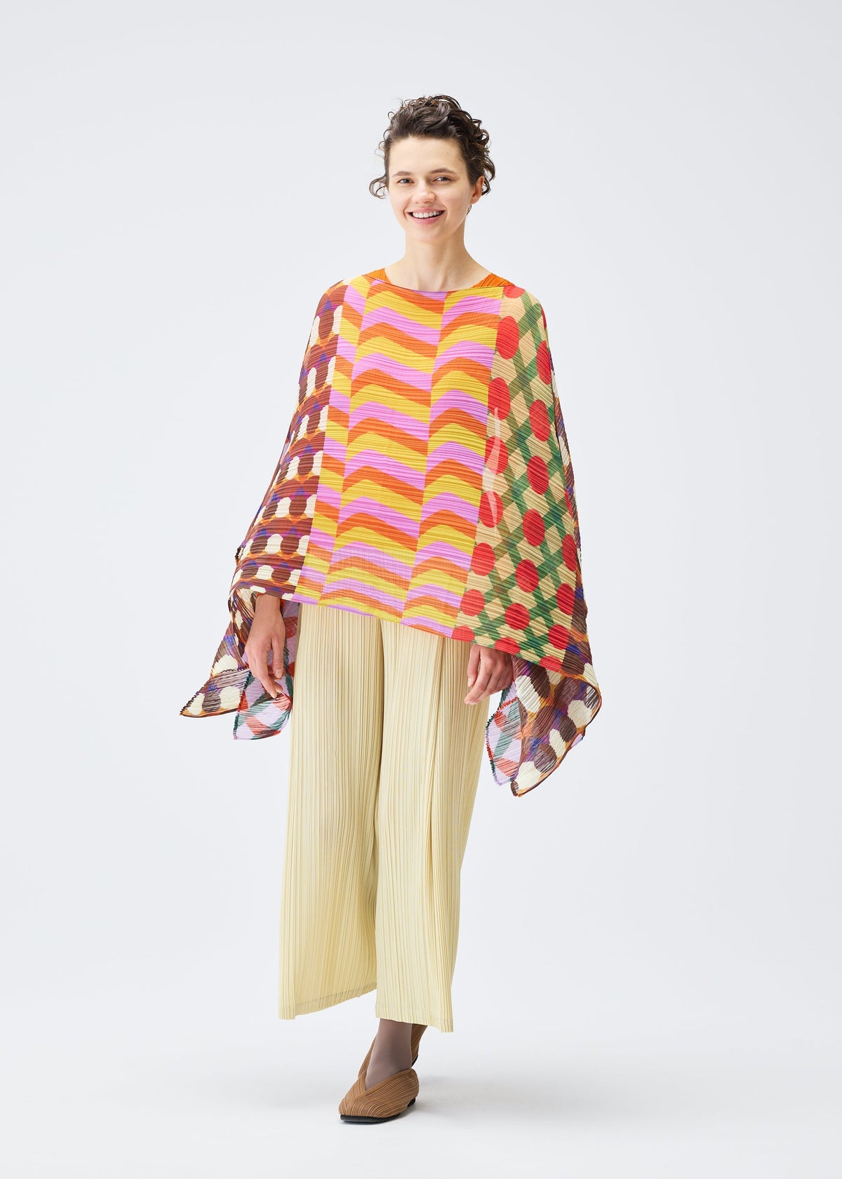 A woman smiles in the PLEATS PLEASE ISSEY MIYAKE HARMONY MADAME-T, a colorful geometric poncho with wavy stripes and dots, over a yellow top and wide light pleated pants. She wears brown shoes and has short curly hair on a white background. view 4 of 4