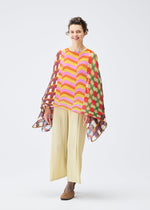 A woman smiles in the PLEATS PLEASE ISSEY MIYAKE HARMONY MADAME-T, a colorful geometric poncho with wavy stripes and dots, over a yellow top and wide light pleated pants. She wears brown shoes and has short curly hair on a white background.