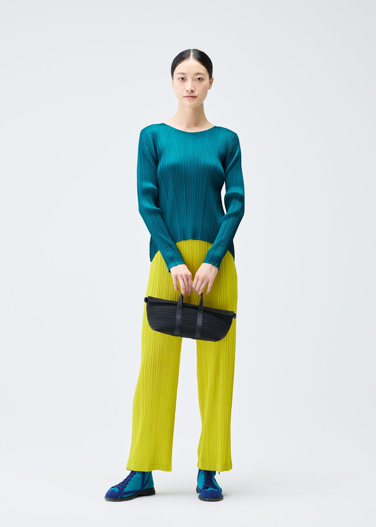 A woman stands against a white background wearing a teal, long-sleeved, horizontally pleated top, bright yellow pleated pants, blue shoes, and holding the PLEATS BOSTON BAG by PLEATS PLEASE ISSEY MIYAKE. view 5 of 6