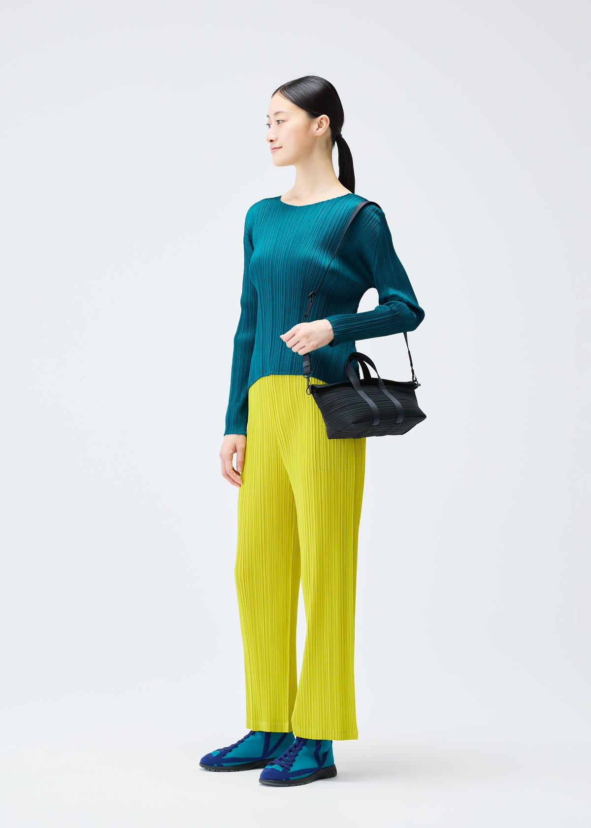 A woman stands against a plain background, wearing a teal pleated top, yellow pleated pants, blue shoes, and carries a PLEATS BOSTON BAG by PLEATS PLEASE ISSEY MIYAKE over her shoulder. She looks to her left. view 6 of 6