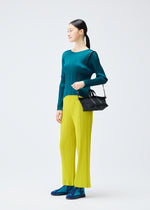 A woman stands against a plain background, wearing a teal pleated top, yellow pleated pants, blue shoes, and carries a PLEATS BOSTON BAG by PLEATS PLEASE ISSEY MIYAKE over her shoulder. She looks to her left.