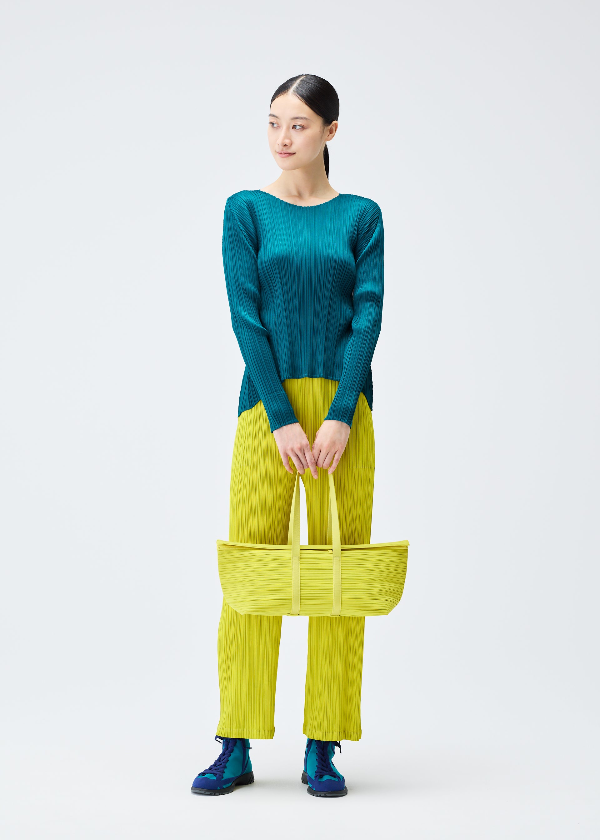 Zoomed view A woman stands against a white background wearing the PLEATS PLEASE ISSEY MIYAKE PP59-AG632 teal long-sleeve top, bright yellow pleated pants, blue shoes, and holding a yellow Boston bag with both hands in front of her.