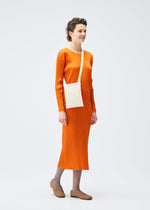 A person stands against a plain white background, smiling to the side. They're wearing a long-sleeved orange ribbed dress, gray tights, tan slip-on shoes, and carrying the PLEATS PLEASE ISSEY MIYAKE CREPE KNIT BAG crossbody.