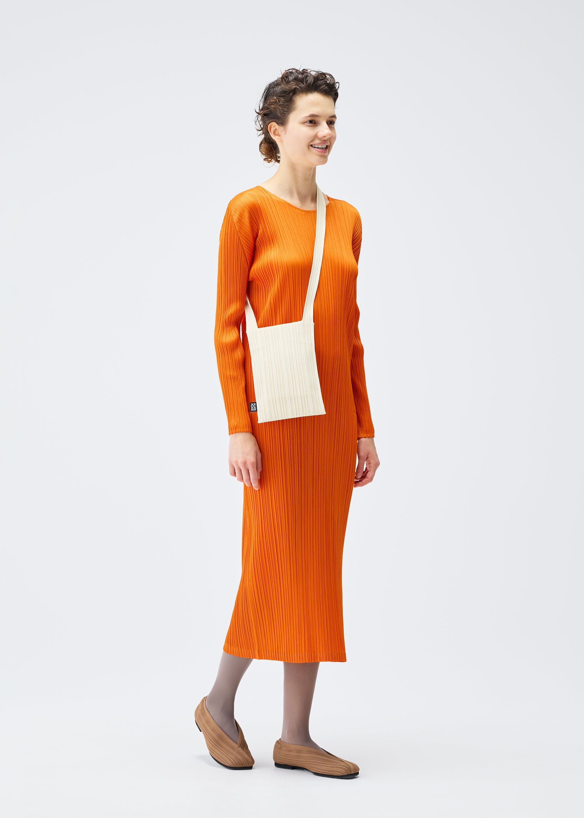 Zoomed view A person stands against a plain white background, smiling to the side. They're wearing a long-sleeved orange ribbed dress, gray tights, tan slip-on shoes, and carrying the PLEATS PLEASE ISSEY MIYAKE CREPE KNIT BAG crossbody.