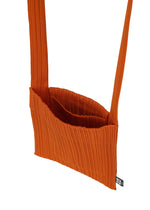 The CREPE KNIT BAG by PLEATS PLEASE ISSEY MIYAKE is a rust orange, ribbed pleated pouch shoulder bag with a wide strap, rectangular body, open top, visible inner pocket, and a small black and white tag at the bottom corner.