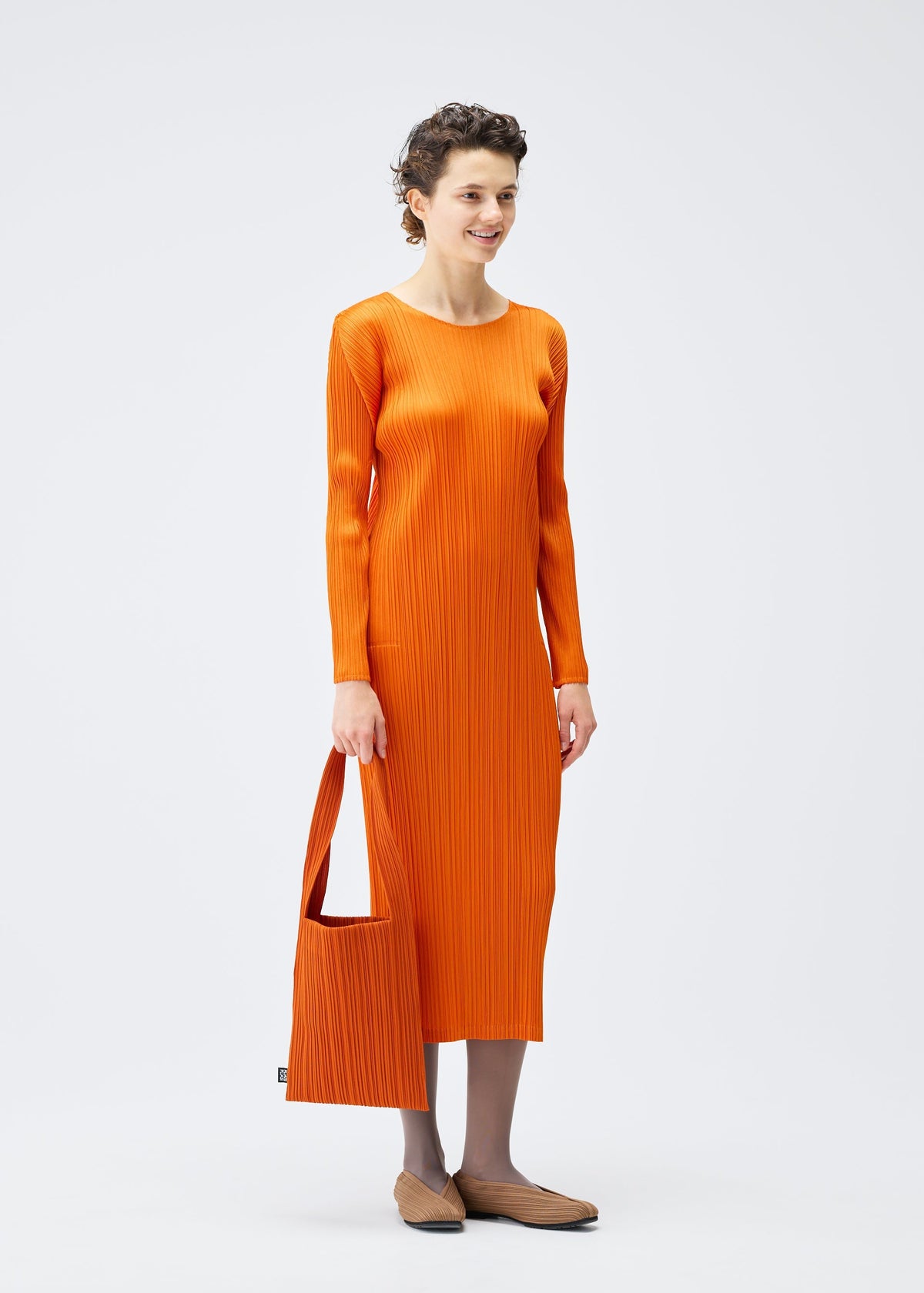 A woman with short, curly hair stands against a plain background, holding the PLEATS PLEASE ISSEY MIYAKE CREPE KNIT BAG in orange. She wears a matching long-sleeved, ankle-length orange dress, brown shoes, and gray tights. view 5 of 5