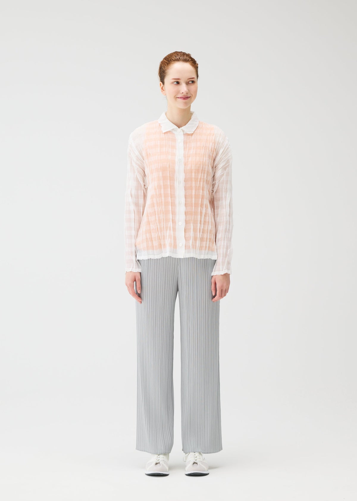 A person faces forward in a sheer, lightweight TWISTED BORDER SHIRT by PLEATS PLEASE ISSEY MIYAKE, paired with light gray pleated pants and white sneakers, set against a plain light background. view 6 of 8