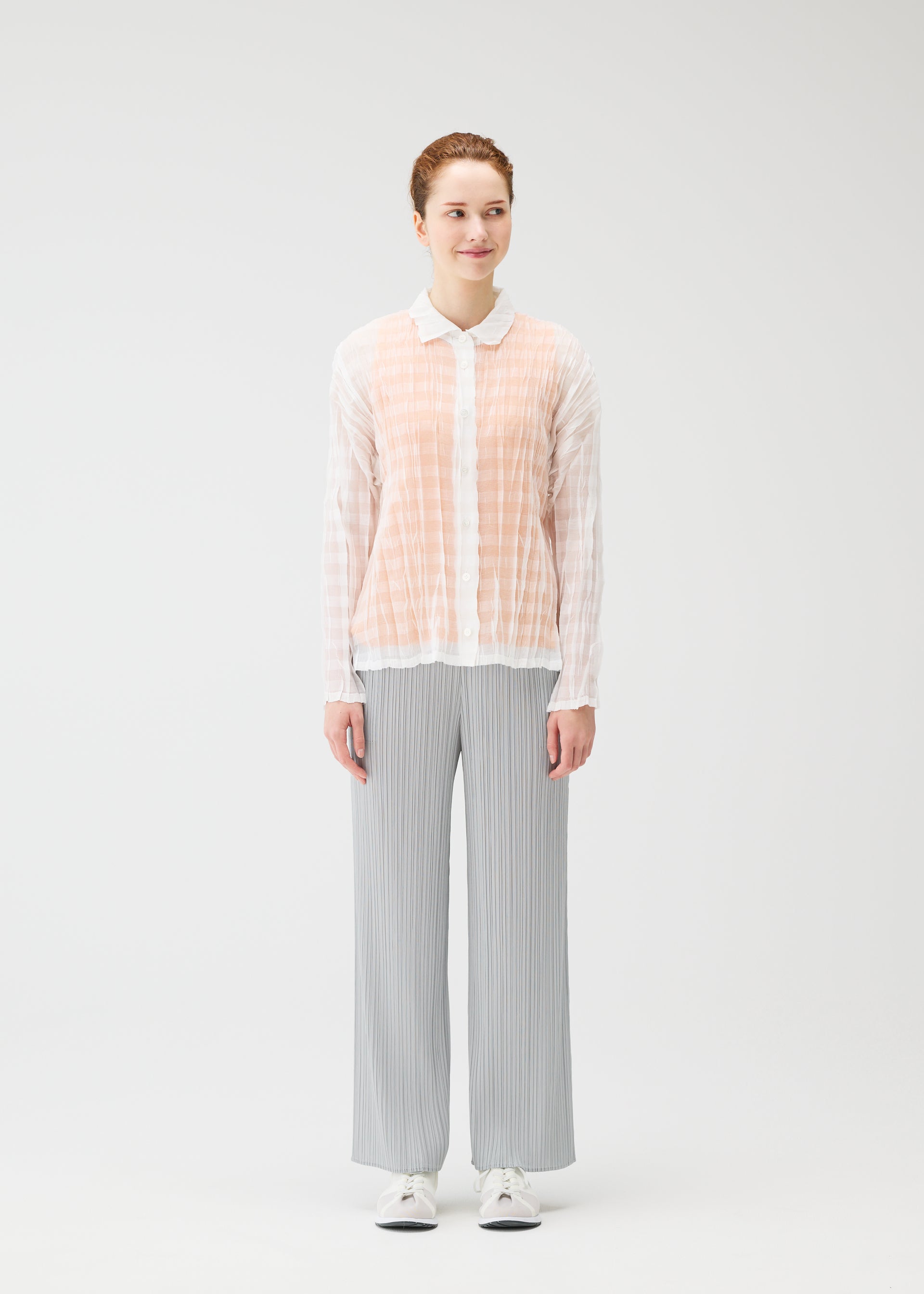 Zoomed view A person faces forward in a sheer, lightweight TWISTED BORDER SHIRT by PLEATS PLEASE ISSEY MIYAKE, paired with light gray pleated pants and white sneakers, set against a plain light background.