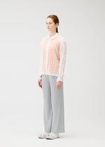 In a white studio, a person faces slightly right, wearing the PLEATS PLEASE ISSEY MIYAKE TWISTED BORDER SHIRT over a white top, paired with light gray pleated pants and white sneakers. Their hair is tied back.