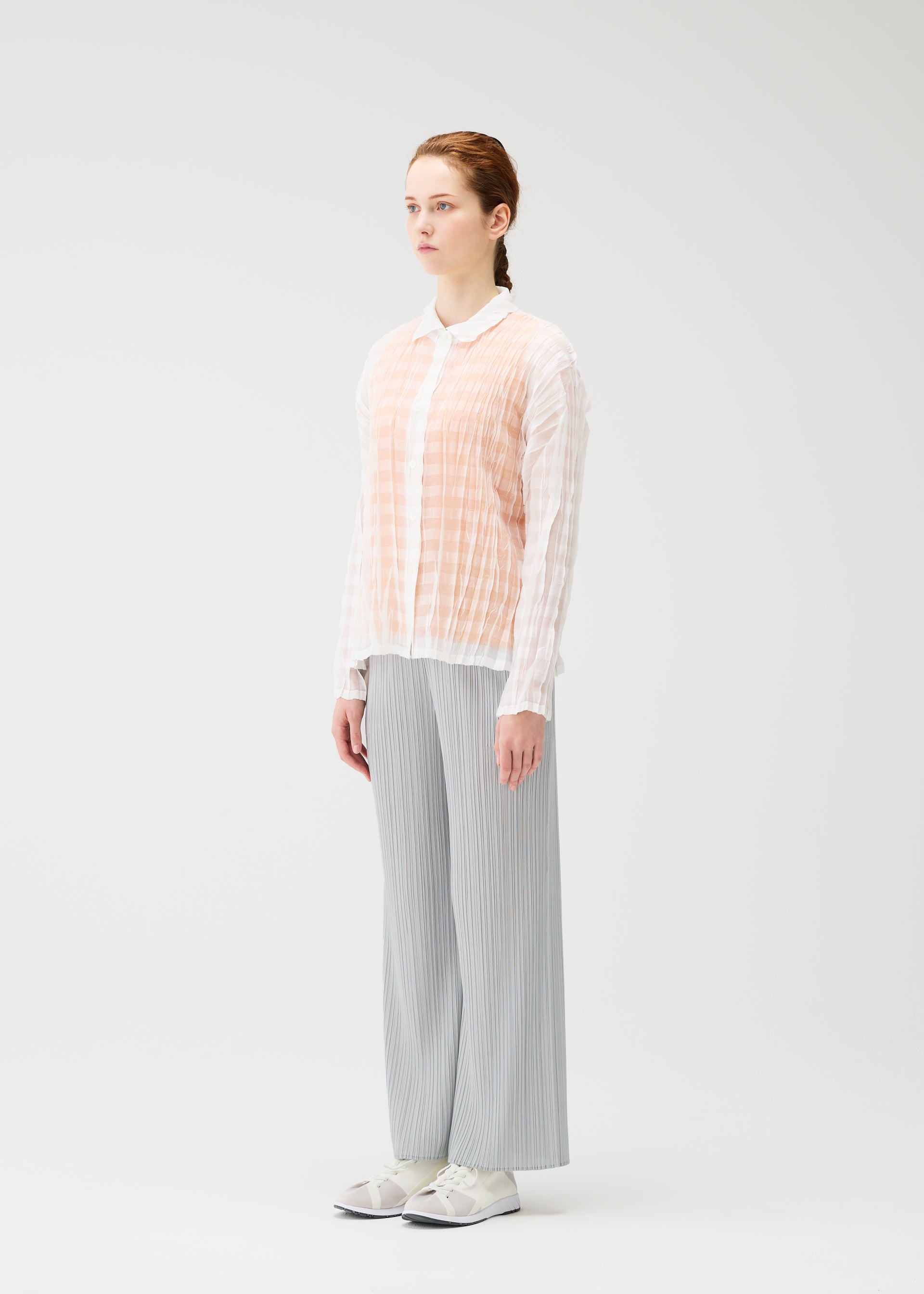 Zoomed view In a white studio, a person faces slightly right, wearing the PLEATS PLEASE ISSEY MIYAKE TWISTED BORDER SHIRT over a white top, paired with light gray pleated pants and white sneakers. Their hair is tied back.