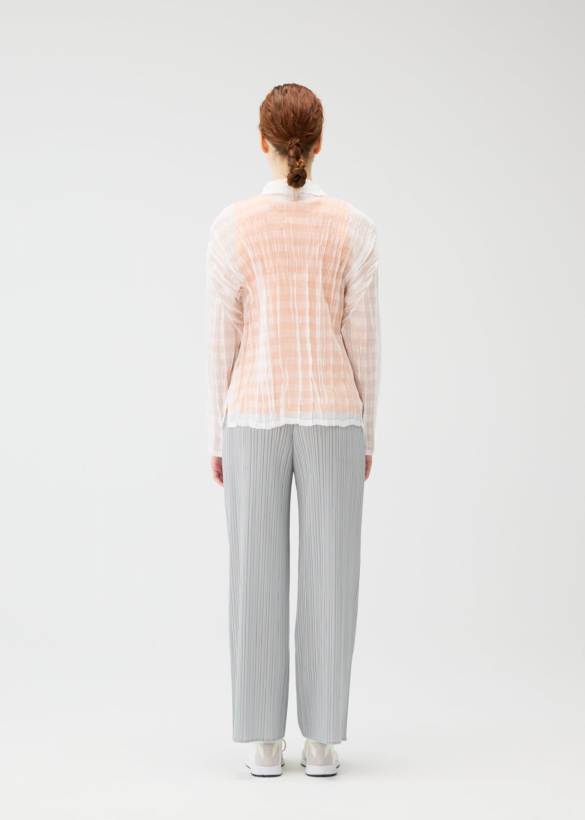 A person with light brown hair tied back stands facing away, wearing the PLEATS PLEASE ISSEY MIYAKE TWISTED BORDER SHIRT under a sheer peach shirt, loose gray pleated pants, and white shoes against a plain light background. view 8 of 8
