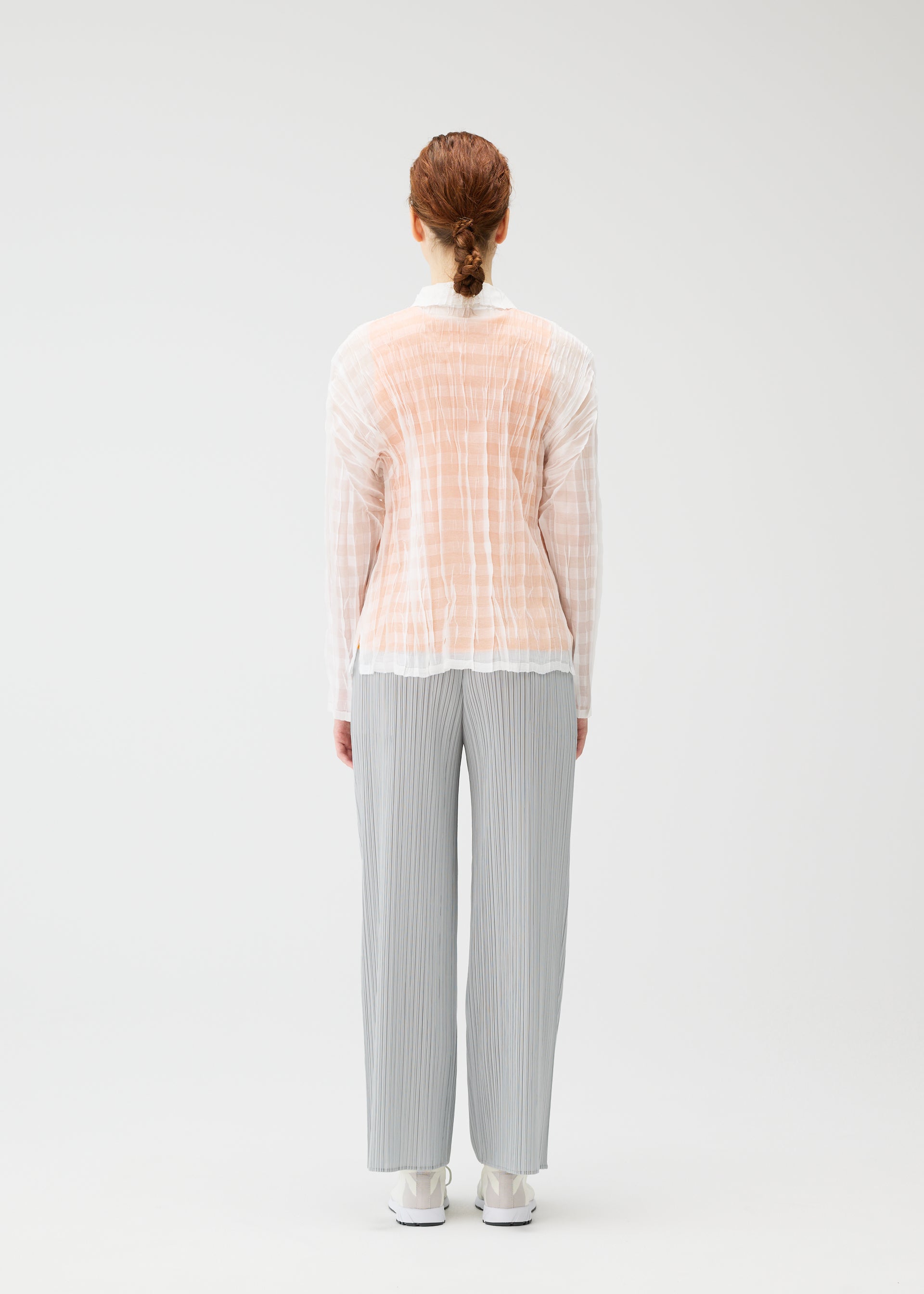 Zoomed view A person with light brown hair tied back stands facing away, wearing the PLEATS PLEASE ISSEY MIYAKE TWISTED BORDER SHIRT under a sheer peach shirt, loose gray pleated pants, and white shoes against a plain light background.
