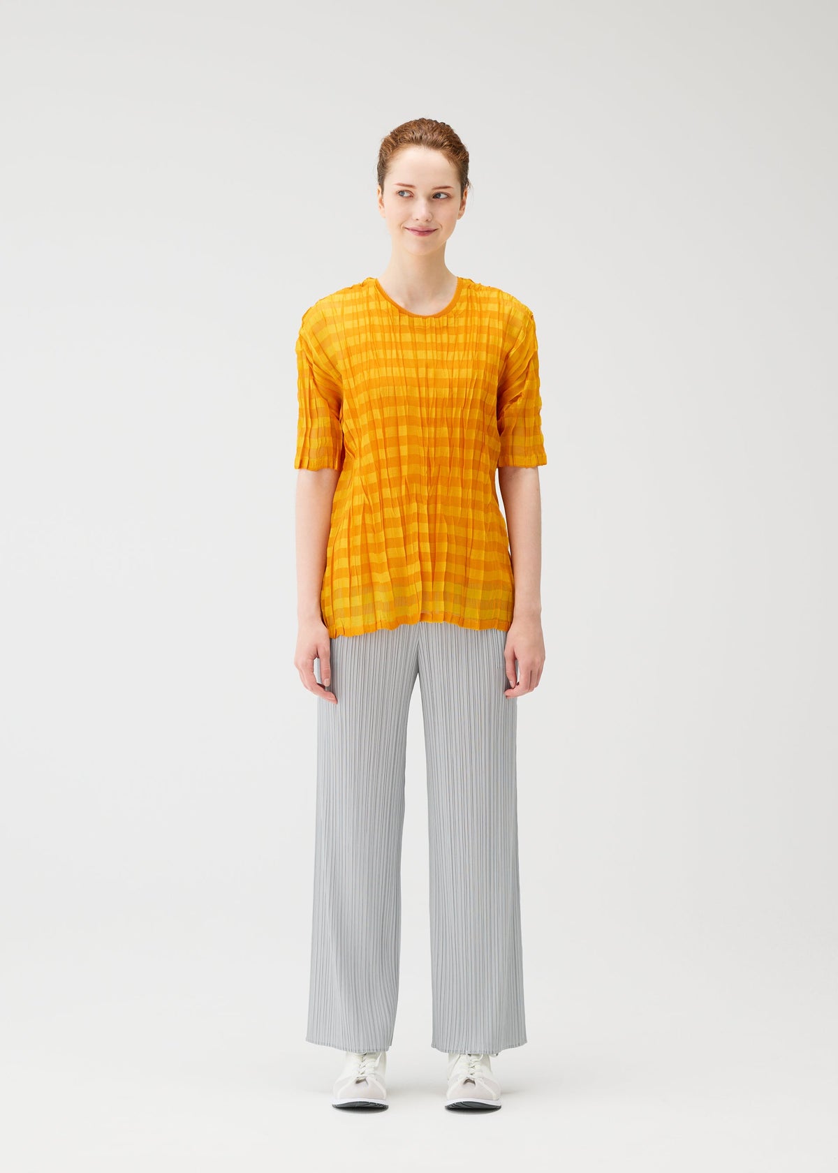 A person faces forward against a white background, wearing the TWISTED BORDER SHIRT by PLEATS PLEASE ISSEY MIYAKE in yellow, paired with light gray wide-leg pants and white sneakers. view 5 of 8