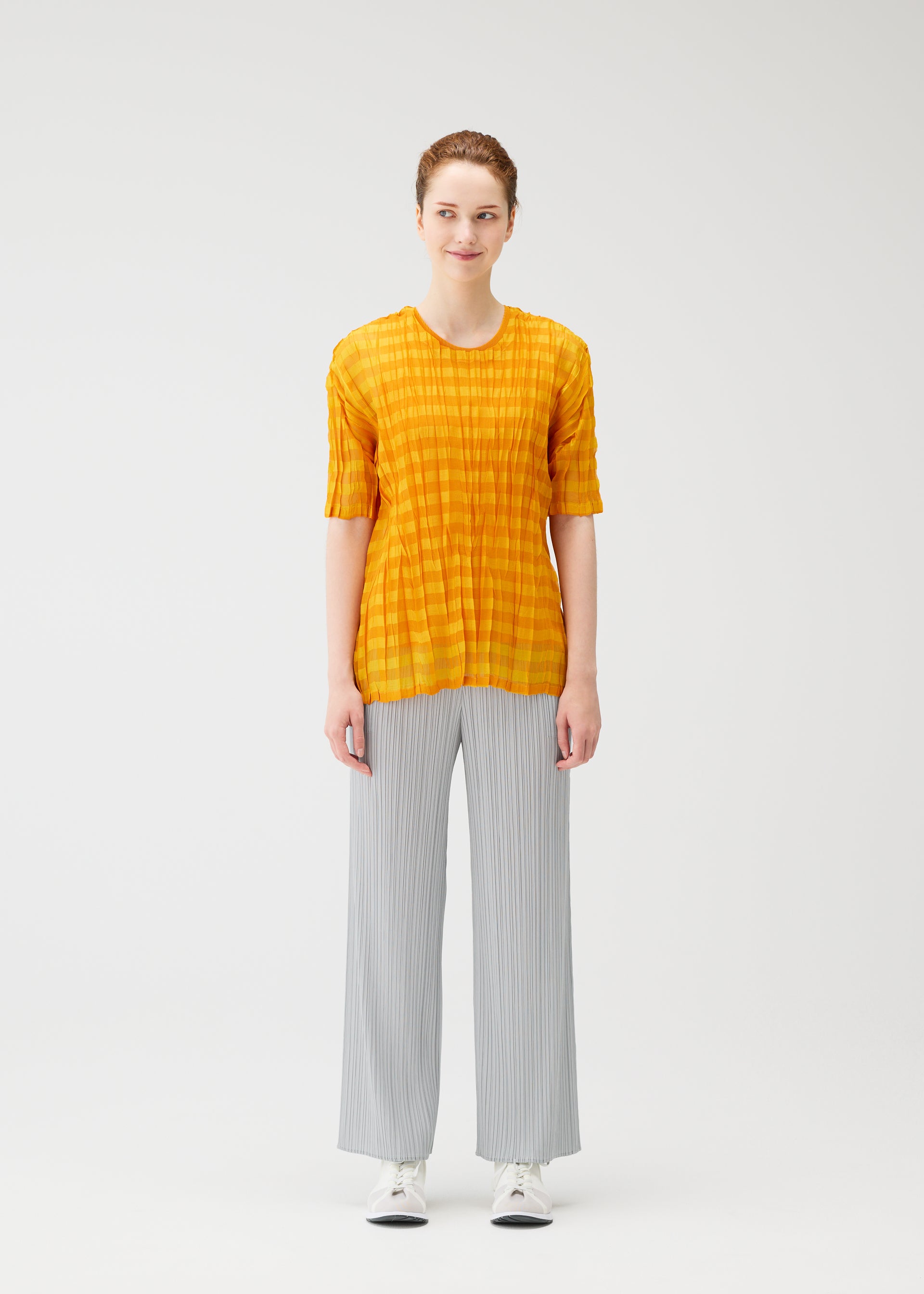 Zoomed view A person faces forward against a white background, wearing the TWISTED BORDER SHIRT by PLEATS PLEASE ISSEY MIYAKE in yellow, paired with light gray wide-leg pants and white sneakers.