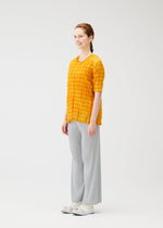 A woman smiles in a bright studio, wearing the TWISTED BORDER SHIRT by PLEATS PLEASE ISSEY MIYAKE, paired with light gray pleated wide-leg pants and white sneakers. Her brown hair is styled in a braid, and she has light skin.