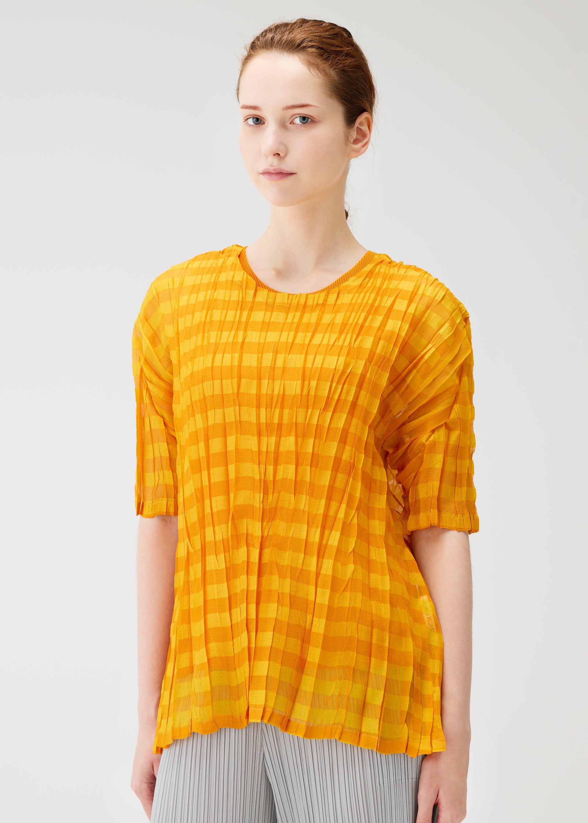 Zoomed view A young woman with fair skin and auburn hair wears the PLEATS PLEASE ISSEY MIYAKE TWISTED BORDER SHIRT, a sheer, pleated yellow-orange short-sleeve top, paired with light gray pleated trousers against a plain light background.