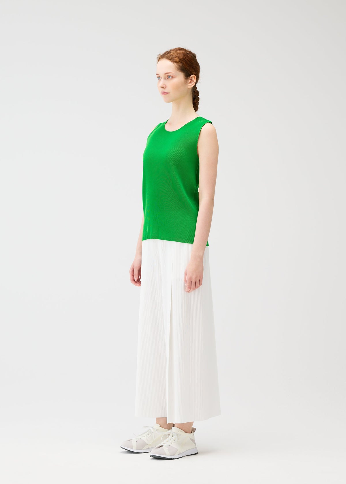 A woman faces left against a plain white background, wearing the sleeveless green "MIST APRIL" pleated tank top by PLEATS PLEASE ISSEY MIYAKE, wide white culotte pants, and white sneakers. view 6 of 8