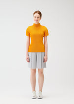 A woman smiles while wearing the MIST APRIL top by PLEATS PLEASE ISSEY MIYAKE— a bright orange, short-sleeved, high-neck piece—paired with light gray knee-length shorts and white sneakers against a white background.