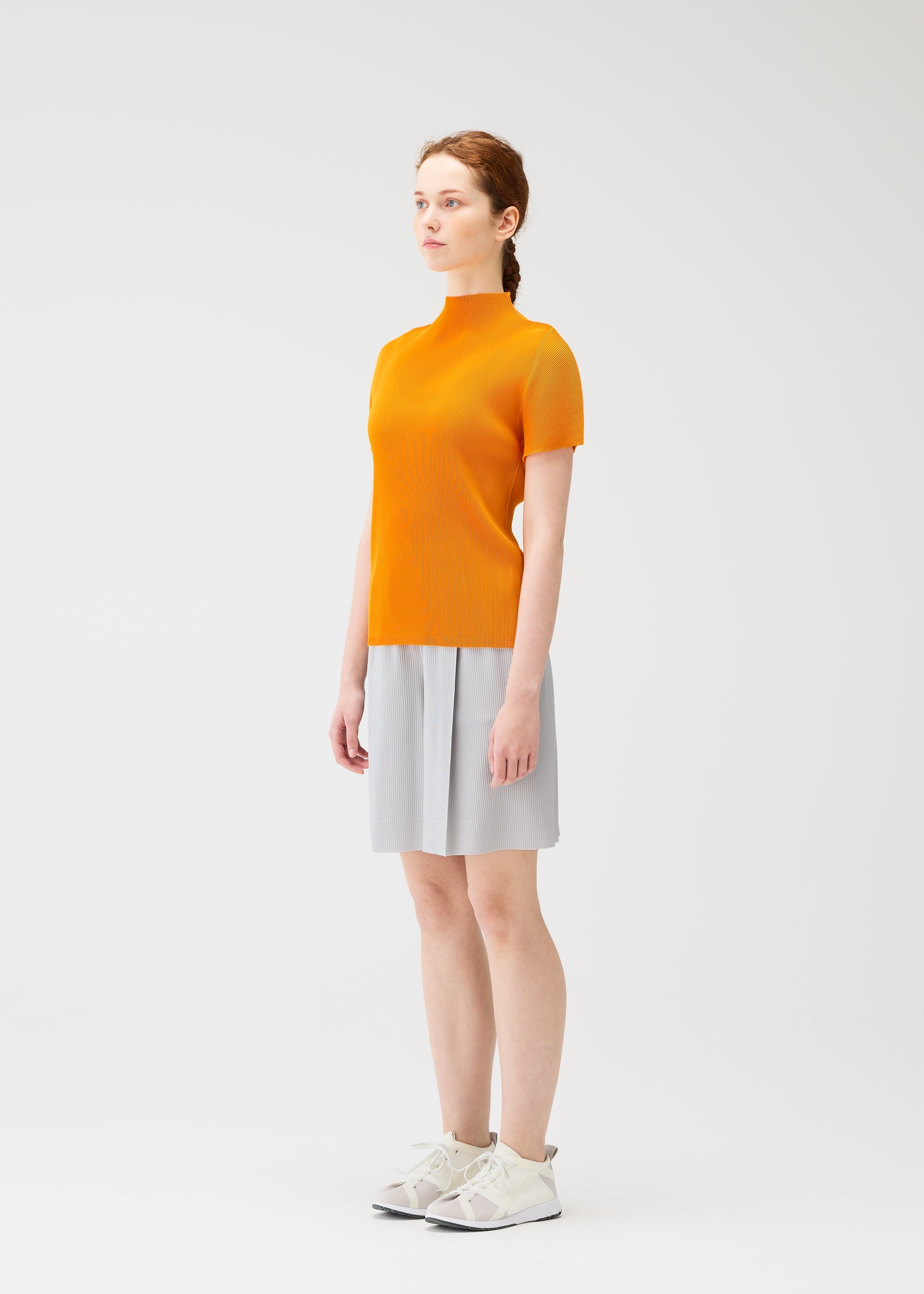 Zoomed view A person with light skin and red braided hair stands against a white background, wearing the MIST APRIL top by PLEATS PLEASE ISSEY MIYAKE, a light gray skirt, and white sneakers, gazing off to the side.