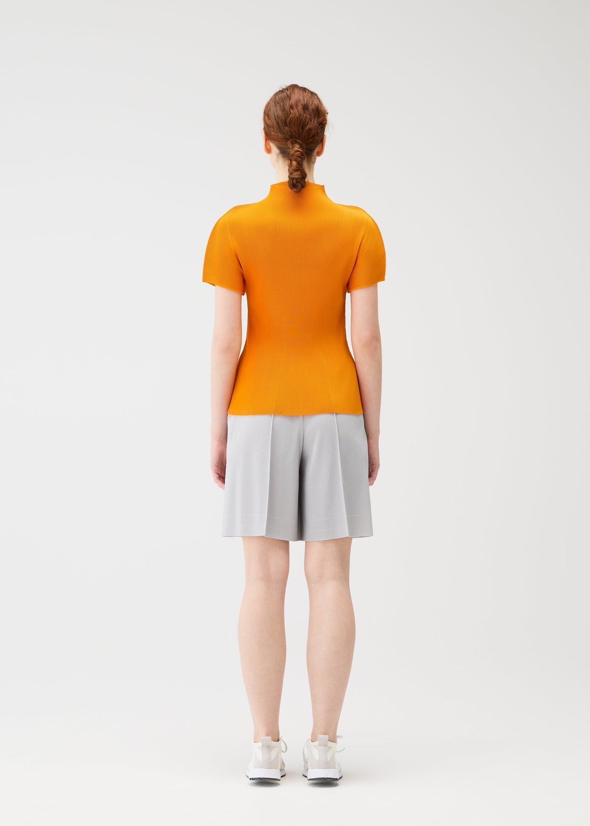A person with red hair in a low bun stands facing away, wearing the bright orange MIST APRIL top by PLEATS PLEASE ISSEY MIYAKE, paired with light gray knee-length shorts and white sneakers, against a plain white background. view 7 of 8