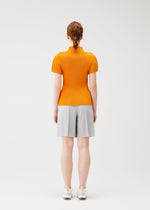 A person with red hair in a low bun stands facing away, wearing the bright orange MIST APRIL top by PLEATS PLEASE ISSEY MIYAKE, paired with light gray knee-length shorts and white sneakers, against a plain white background.