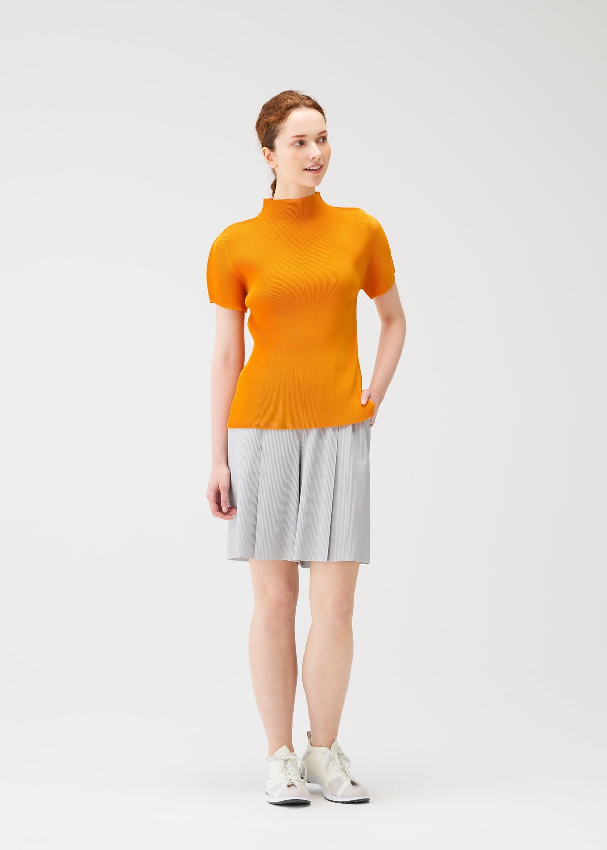 A woman with light skin and brown hair tied back smiles slightly, posing in the MIST APRIL top by PLEATS PLEASE ISSEY MIYAKE—an orange high-neck, short-sleeved top paired with a light gray pleated skirt and white sneakers. view 8 of 8