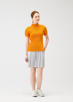 A woman with light skin and brown hair tied back smiles slightly, posing in the MIST APRIL top by PLEATS PLEASE ISSEY MIYAKE—an orange high-neck, short-sleeved top paired with a light gray pleated skirt and white sneakers.